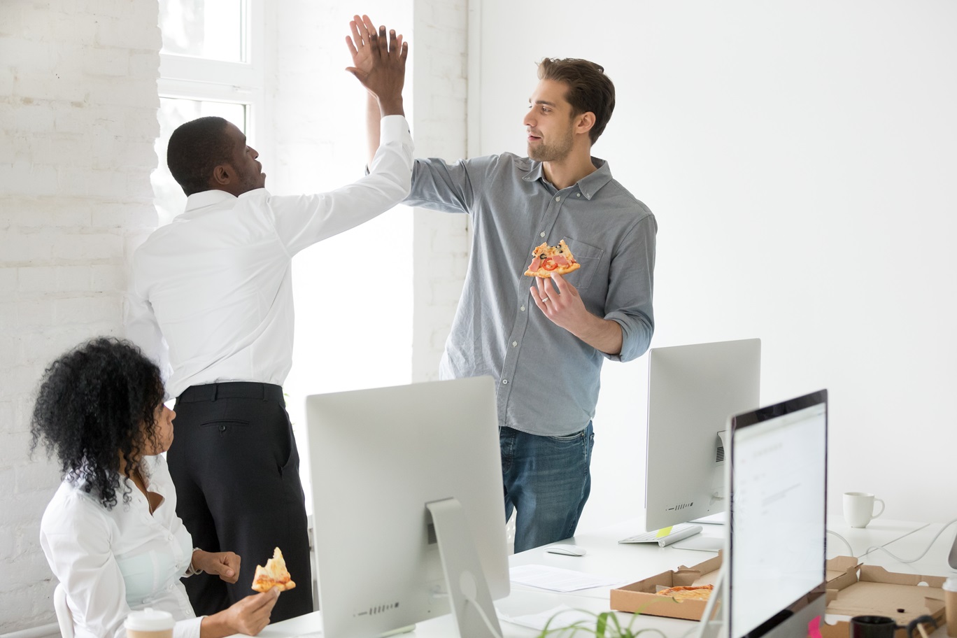 Smiling multiracial coworkers giving high five eating pizza together at office break, happy diverse male colleagues join hands celebrating success achievement, friendship and good relations concept