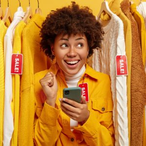 Sale, fashion, discounts and online shopping concept. Overjoyed dark skinned woman chooses clothes in clothing store, rejoices big sales, holds mobile phone for making payment via special app