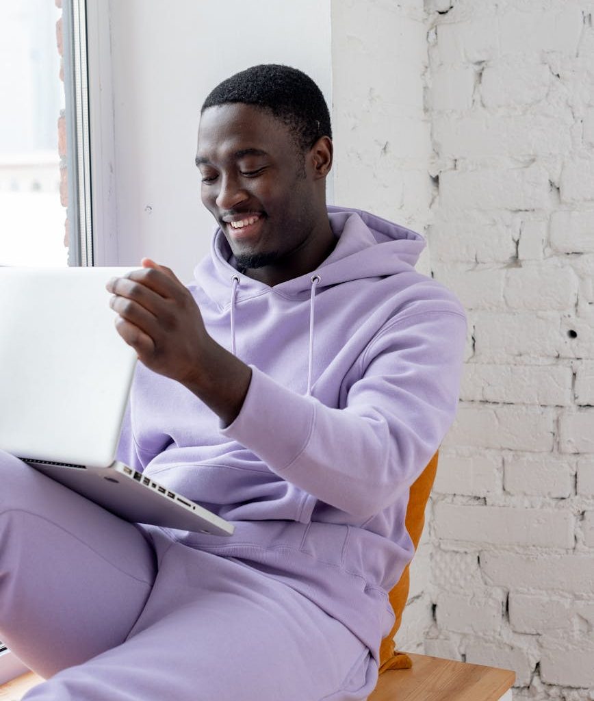 black cheerful man using laptop on windowsill